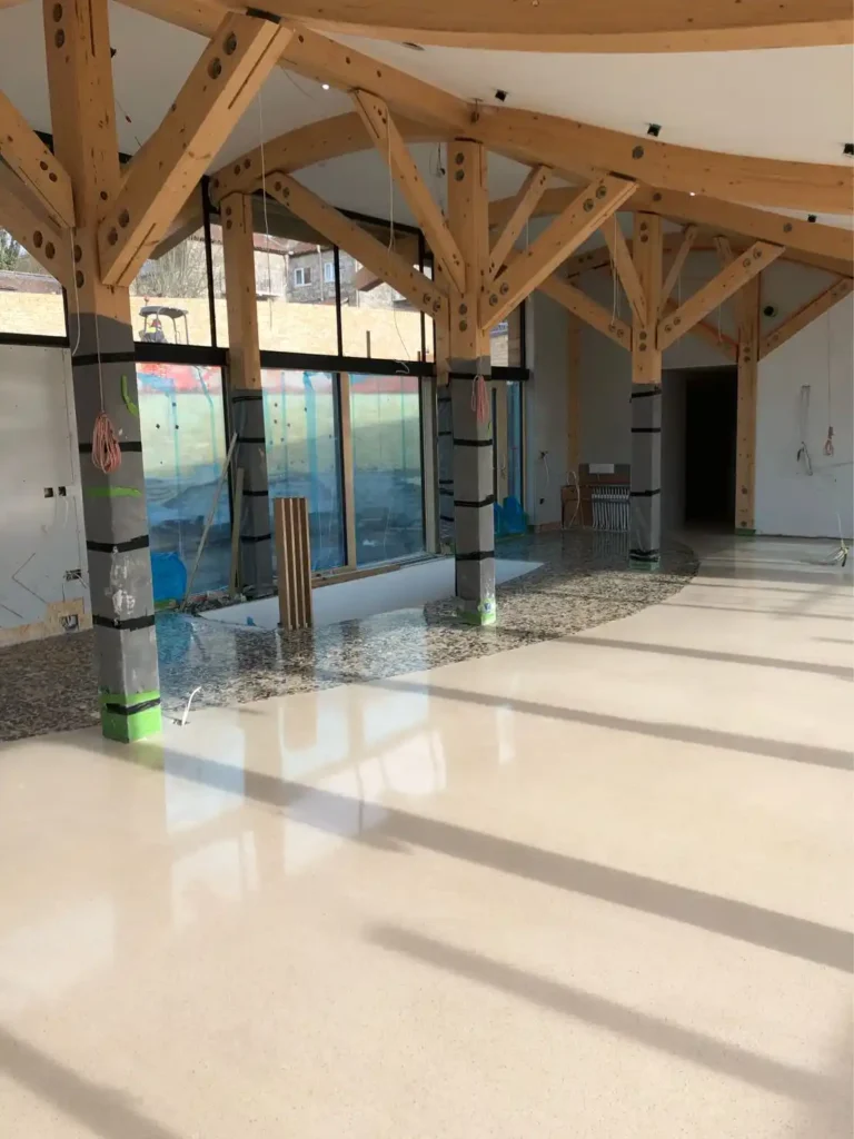 Espacio en construcción con estructura de madera, suelo de terrazo beige y grandes ventanales.