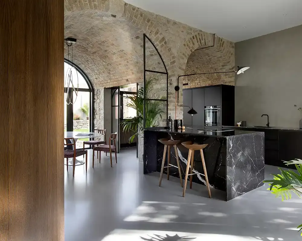 Cocina moderna con paredes de piedra, techo abovedado y una isla de mármol negro, con mobiliario minimalista.