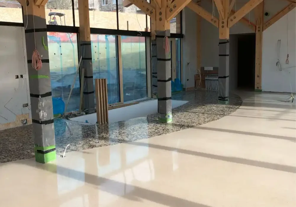 Espacio en construcción con estructura de madera, suelo de terrazo beige y grandes ventanales.