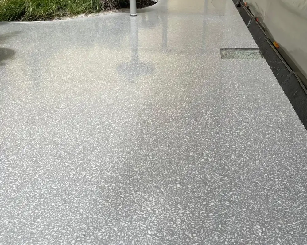 Suelo de terrazzo gris en una zona exterior con plantas y una estructura en construcción al fondo.